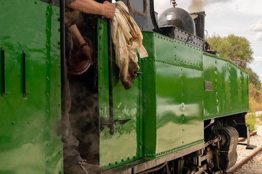 A green steam locomotive with a person standing at the open door, a steam pipe, and a track. 1