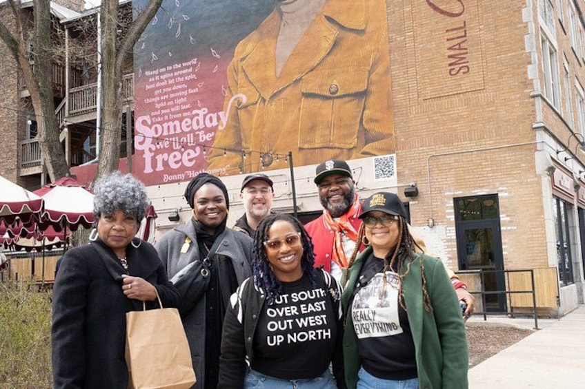 A group of people standing in front of a building with a large mural and a sign that reads 'Someday We'll All Be Free.' 4