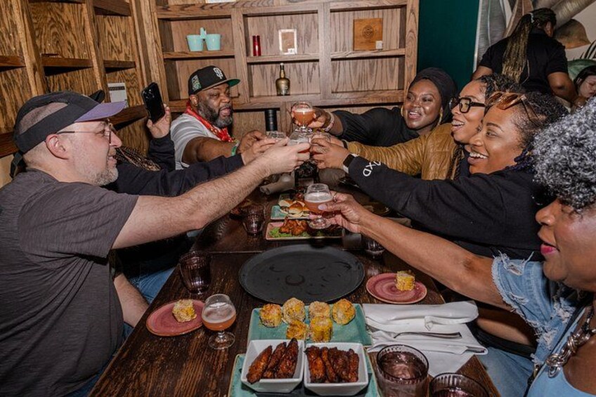 A group of people toasting at a wooden table with food and drinks. 1