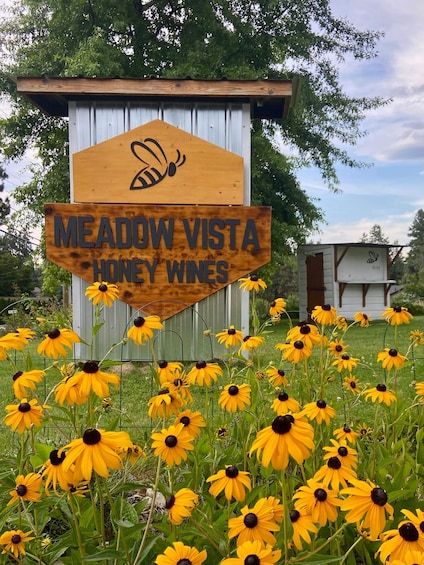 A wooden sign with a bee and text for Meadow Vista Honey Wines, surrounded by yellow flowers. 3