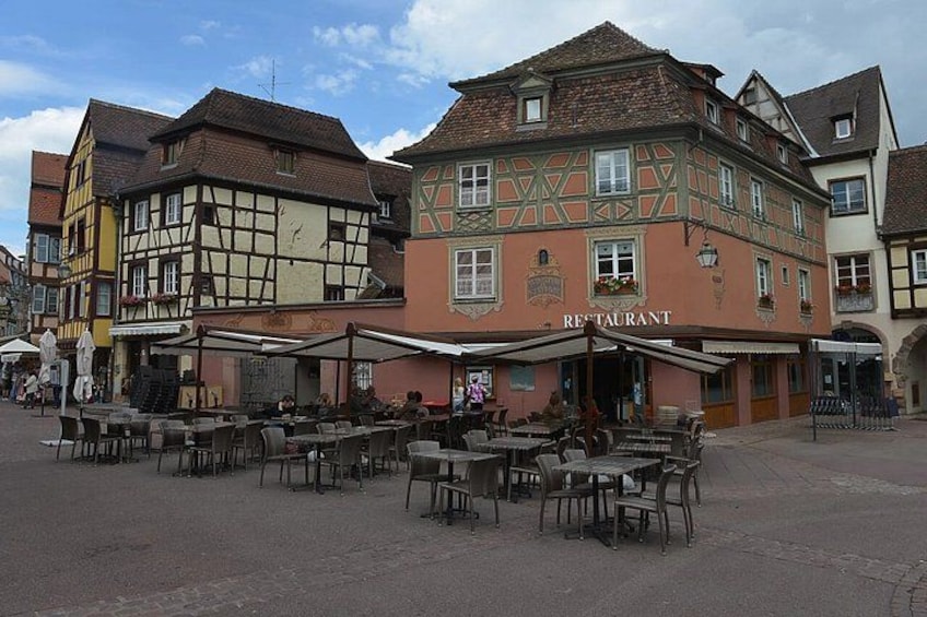 A European town square with traditional half-timbered buildings, outdoor seating at a restaurant, and a cobblestone street. 5