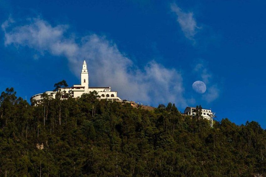 A white church with a bell tower on a hill, surrounded by dense forest, under a clear blue sky with a visible moon. 1