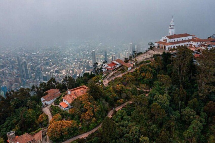 A mountainous landscape with a prominent white building with a tower, surrounded by dense greenery and a cityscape in the background. 5
