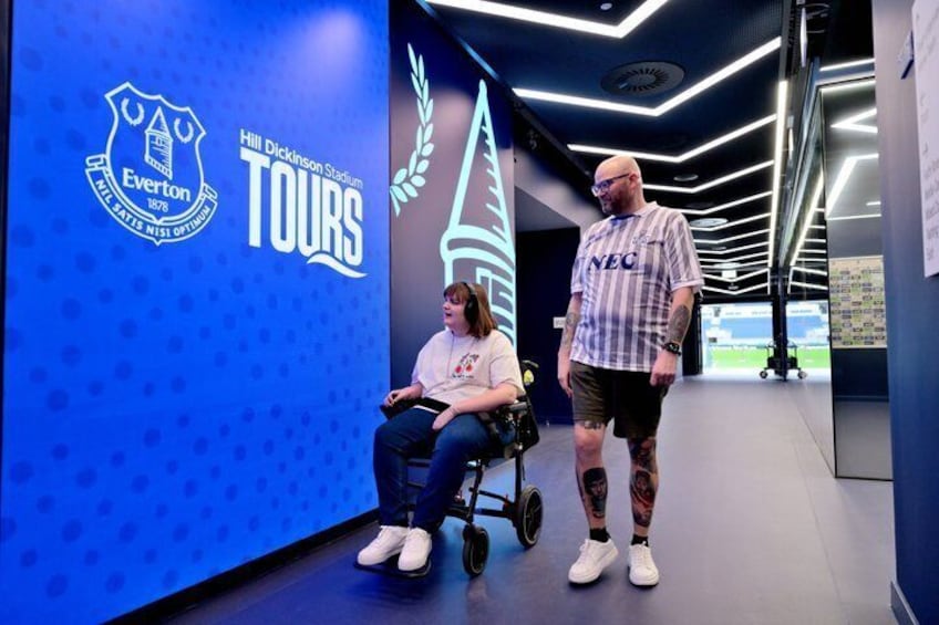 A person in a wheelchair, another standing, and a wall with Everton and Hill Dickinson Stadium logos. 3