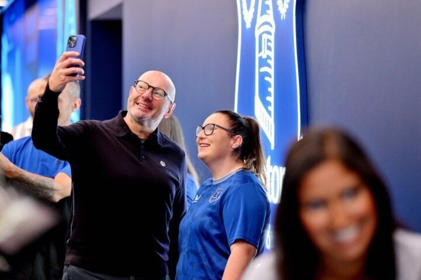 Two individuals taking a selfie with a blue backdrop and a logo. 2