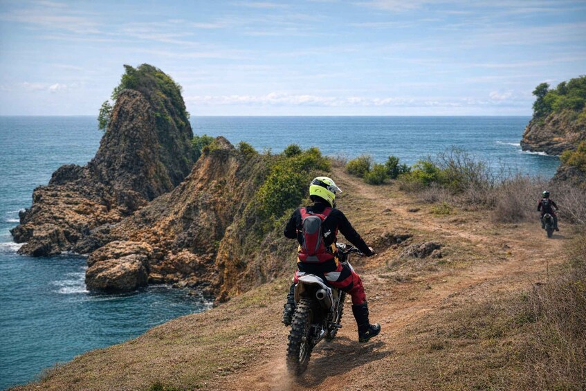 A person on a dirt bike on a coastal trail with rocky cliffs and the ocean in the background. 1
