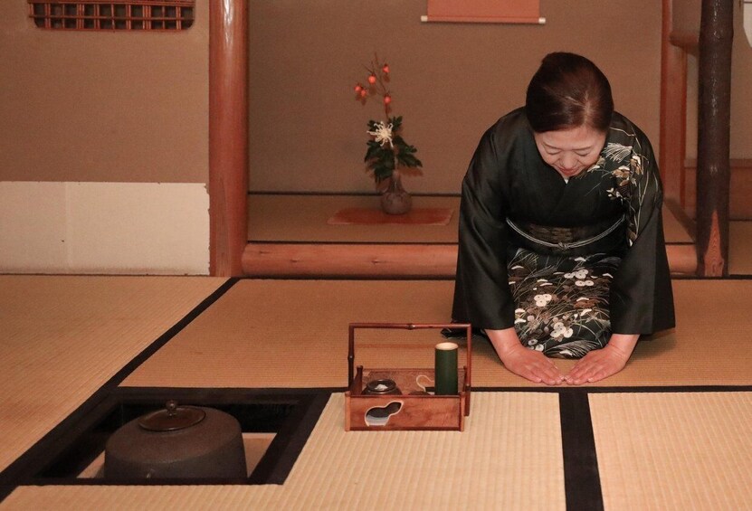 A person in traditional attire kneeling on tatami mat flooring, with a small wooden box and a candle in front. 2