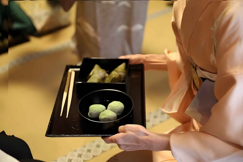 A person holding a tray with a black box containing green dumplings and a bowl of green balls. 5