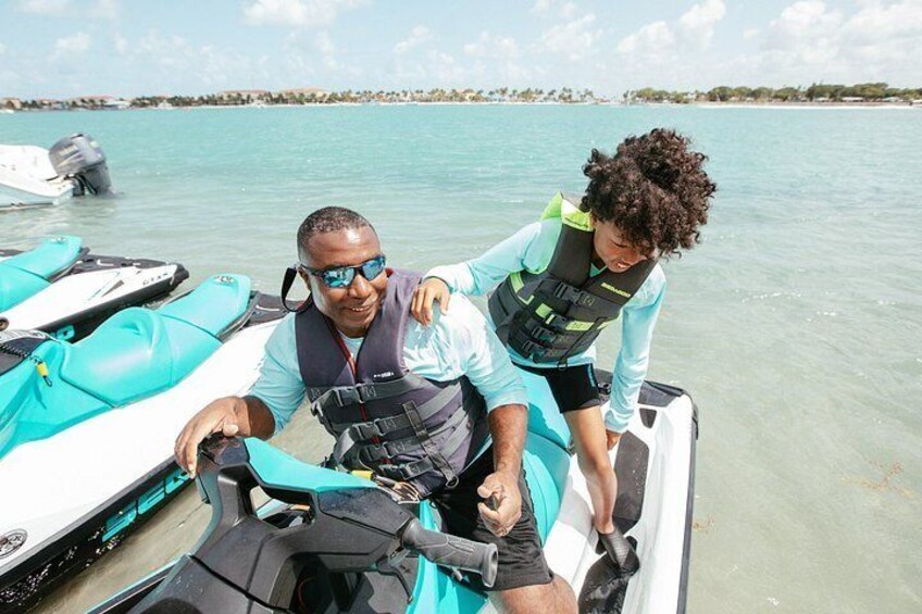 A person in a life jacket on a jet ski, another person assisting, and a calm body of water in the background. 3