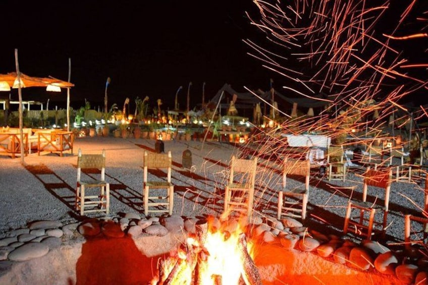 A nighttime outdoor setting with a fire, metal chairs, and a structure with a roof. 3