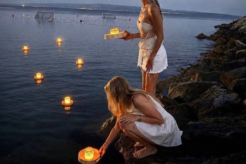 A woman in a white dress by the water, holding a candle, with others around. 1