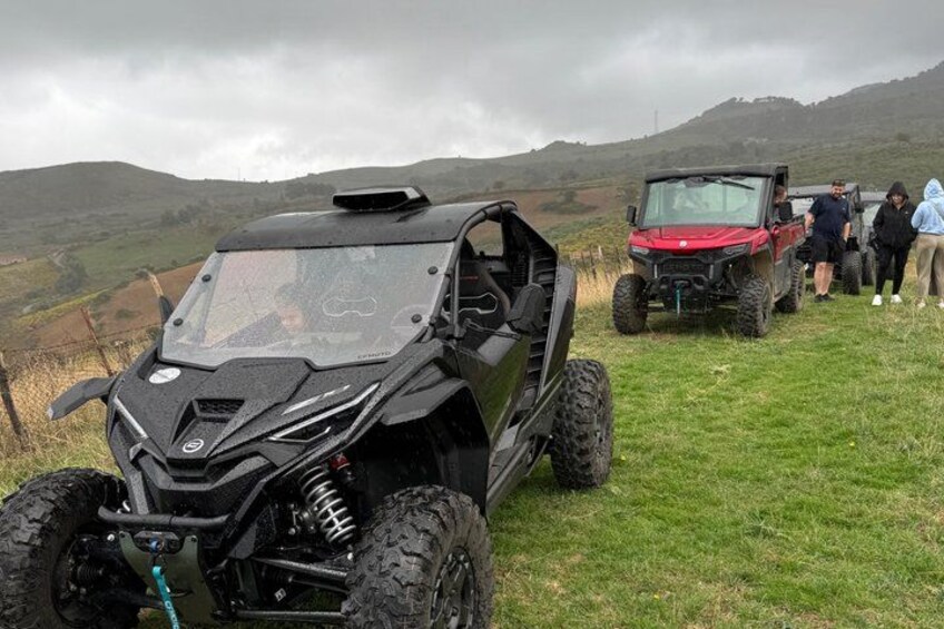 A black off-road vehicle parked on a grassy field with a red vehicle and people in the background. 5
