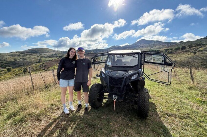 Two people standing next to a black ATV in a grassy field with hills in the background. 3
