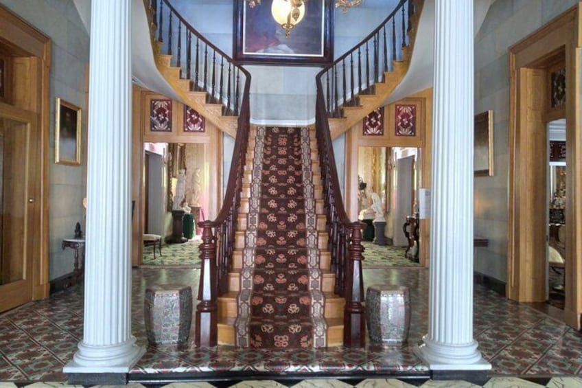 A grand staircase with ornate railings and patterned carpet, flanked by columns and leading to a landing with a chandelier. 3