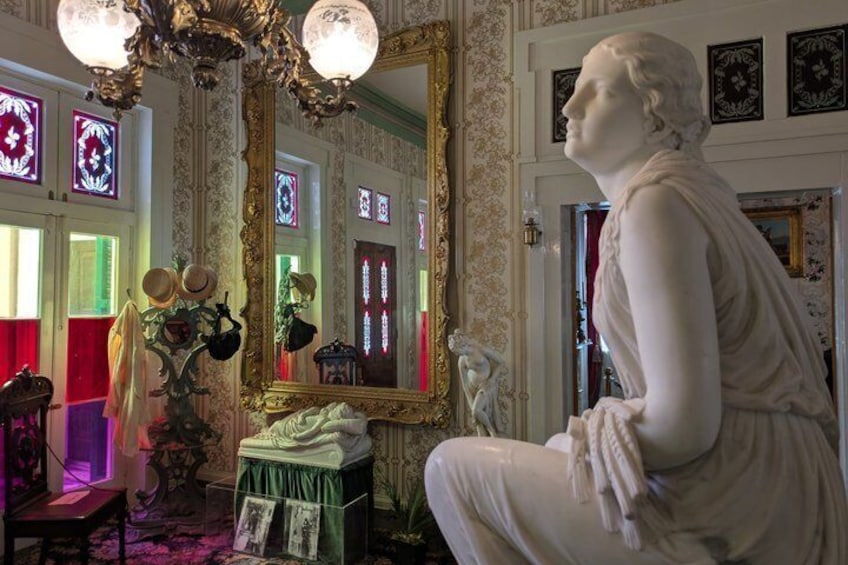 A statue in a room with ornate wallpaper, a large mirror, and stained glass windows. 4