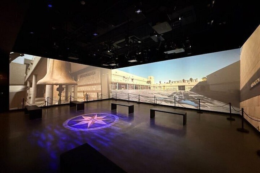 An indoor exhibition space with a large projection screen displaying a historical scene, a star-shaped light on the floor, and a wooden wall with the text 'HISTORY'. 2