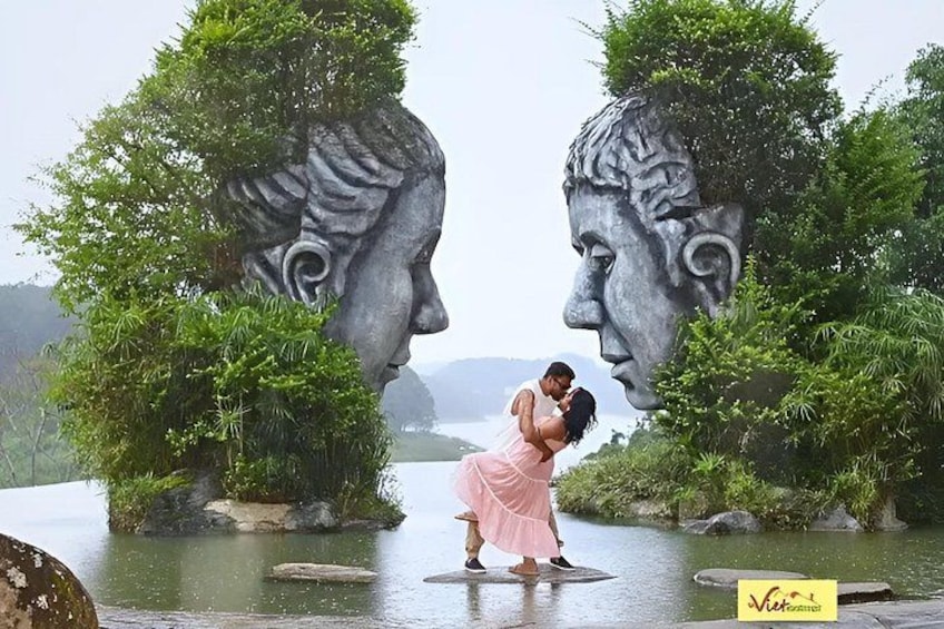 A couple embracing in a garden with large sculpted heads and lush greenery. 1