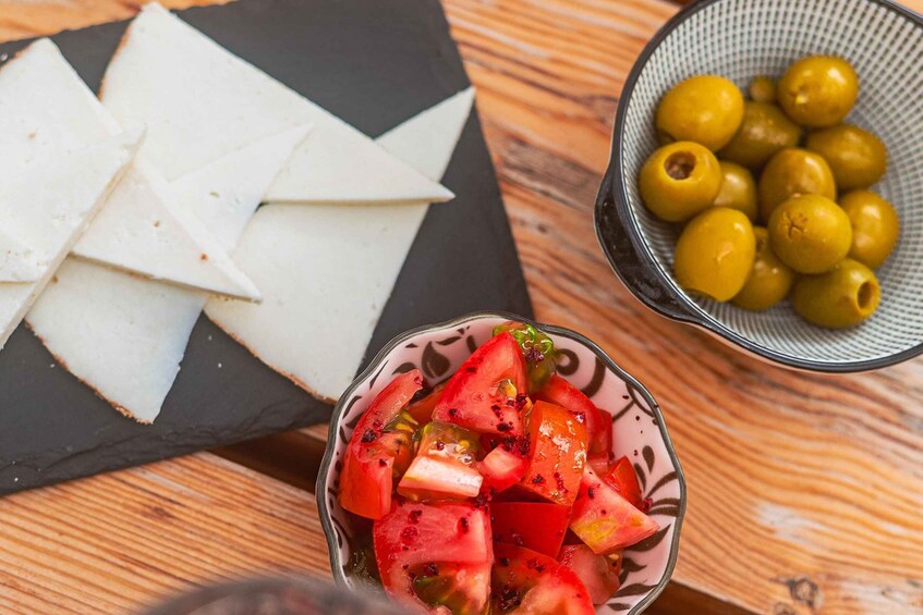 Sliced white cheese, a bowl of diced tomatoes, and a bowl of green olives on a wooden table. 2