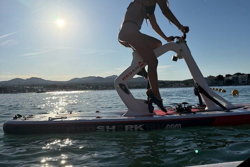 A person riding a stationary bike on a water platform with a cityscape and mountains in the background. 2