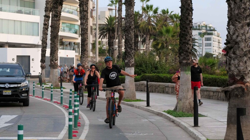Ciclistas en una acera urbana, con palmeras y edificios modernos al fondo. 1