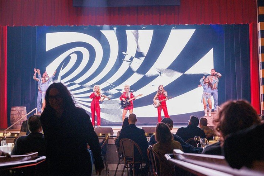A stage with performers in red outfits, a large screen displaying a spiral pattern, and an audience seated at tables. 5