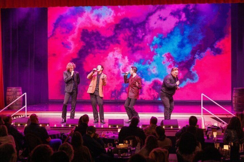 A stage with four people performing, a large screen displaying abstract art, and an audience seated in front. 5