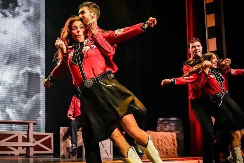 A stage with performers in red jackets and black skirts, a large screen in the background, and a wooden structure. 4