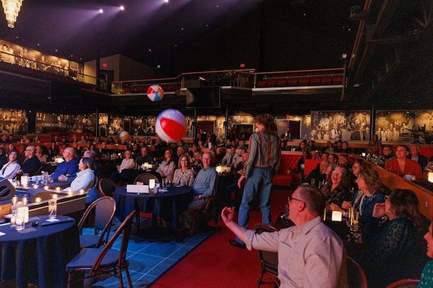 A crowded indoor event with tables, chairs, and a person standing in the center. 2