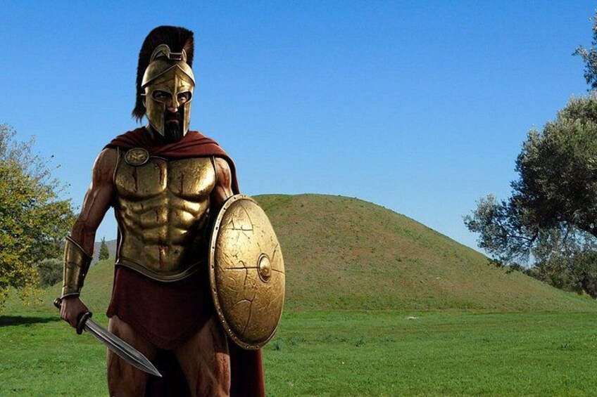 A bronze statue of a warrior holding a sword and shield, standing on a grassy hill with trees in the background. 1