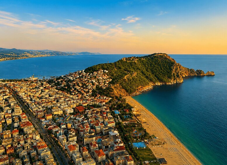 An aerial view of a coastal city with dense urban areas, a prominent hill with ruins, and a clear blue sea. 1