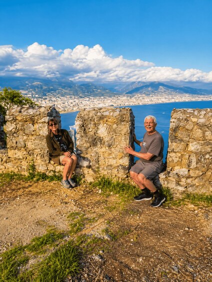 Two people sitting on stone walls overlooking a coastal city with mountains in the background. 4