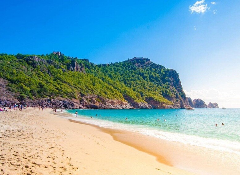 A sandy beach with clear turquoise water, a green hill with a castle, and rocky outcrops. 5