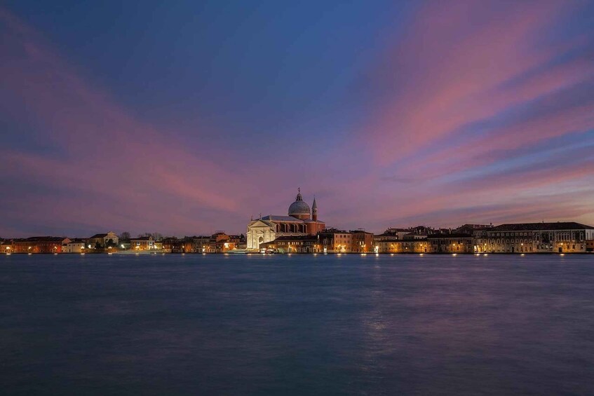 A waterfront cityscape at dusk with illuminated buildings and a prominent domed structure. 1