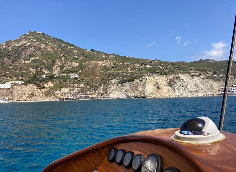 A boat's wooden bow with a radar dome and a rocky coastline with buildings in the background. 4
