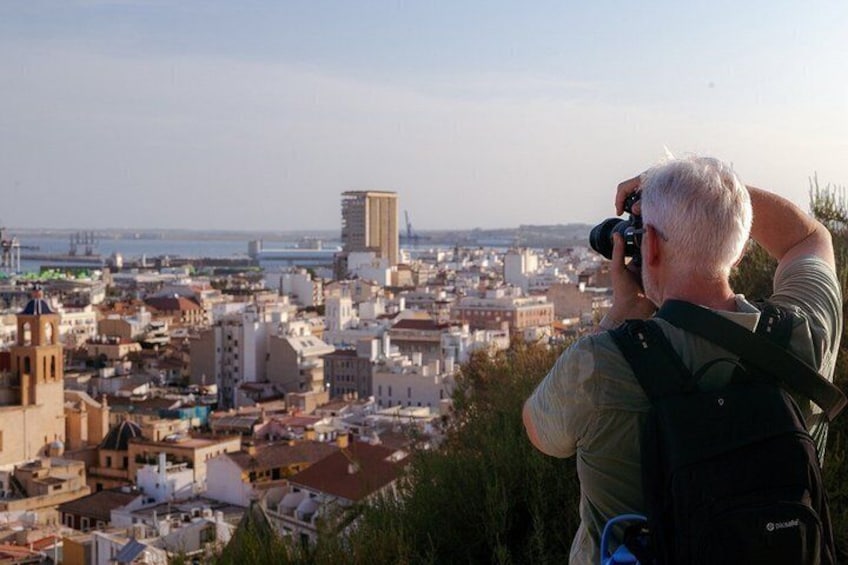 A person taking a photograph of a cityscape with buildings and a distant view of the sea. 2