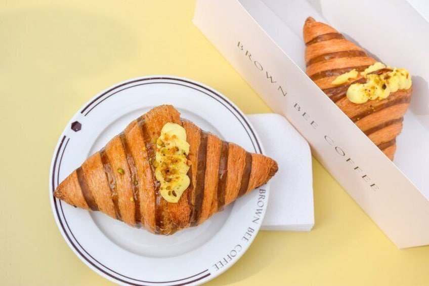 A croissant with a pat of butter on a plate, and another croissant in a box labeled 'BROWN BEE COFFEE'. 1