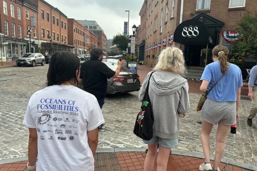 People walking on a cobblestone street, a black car parked on the side, and a building with the number 888 on its facade. 4