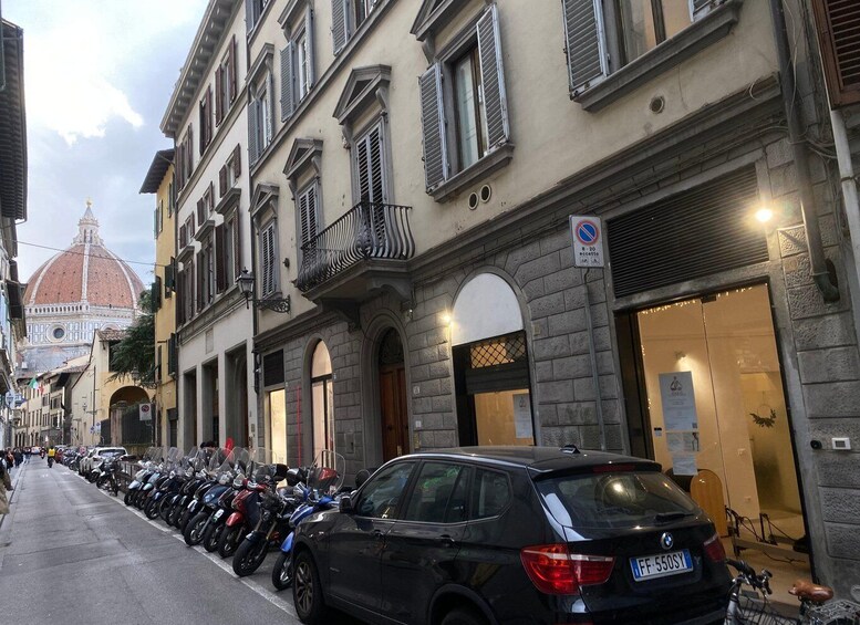 A row of parked motorcycles and a black car on a city street with historic buildings and a dome in the background. 2