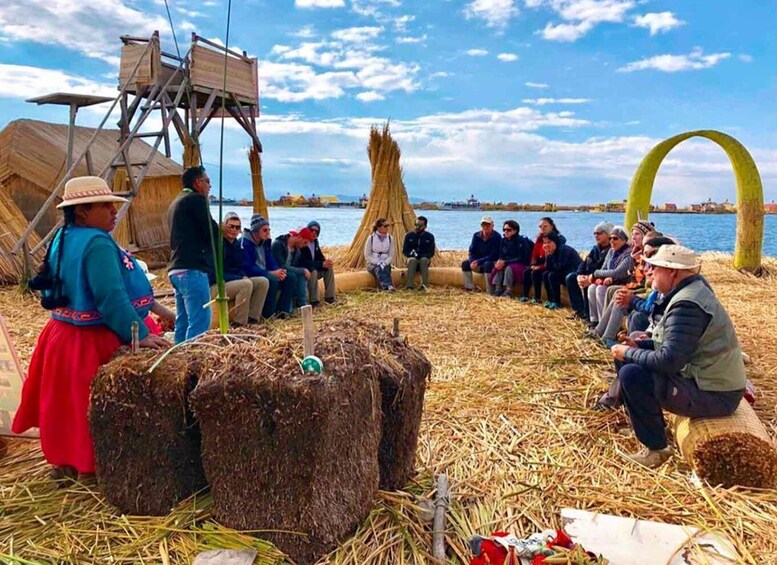 A group of people seated on straw-covered ground, with a wooden structure and a large arch in the background. 3