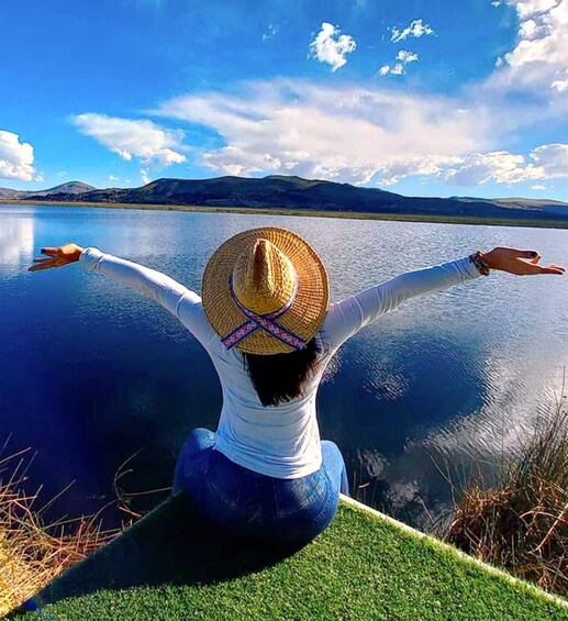 A person sitting by a calm lake with arms outstretched, wearing a straw hat and a white top. 4