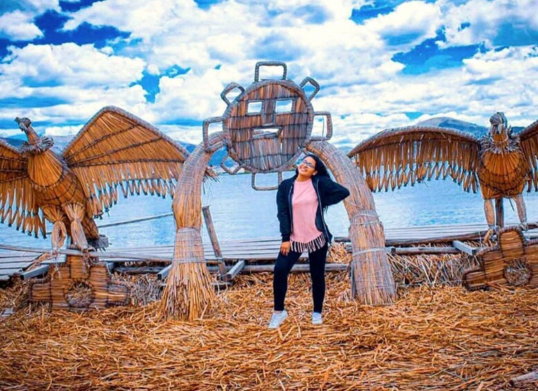 A person standing in front of a large, wooden, mechanical sculpture with wings, surrounded by straw on a beach with a body of water and a cloudy sky in the background. 1