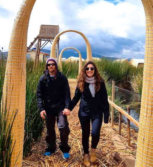 Two people walking on a straw-covered path with a large circular structure in the background. 2