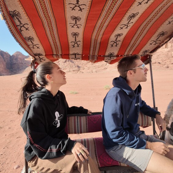 Two people sitting under a patterned umbrella in a desert landscape. 5