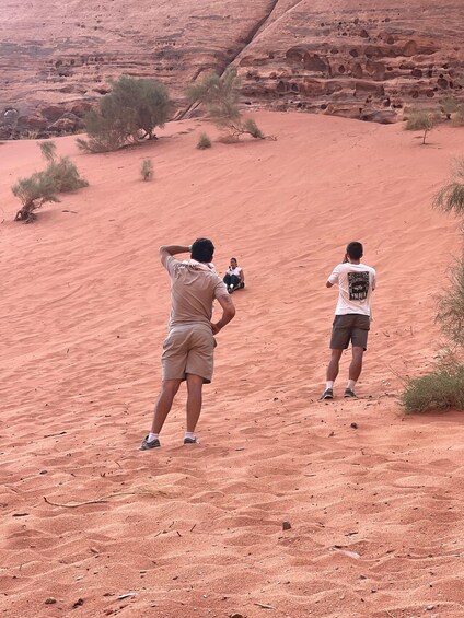Two people taking photographs in a desert landscape with red sand and sparse vegetation. 2