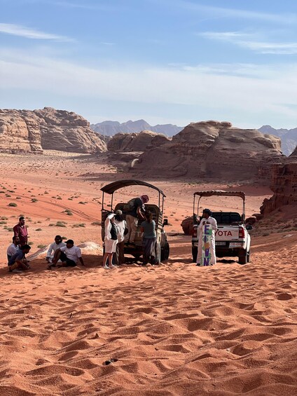 A group of people around a vehicle in a desert landscape with rocky formations and mountains in the background. 1
