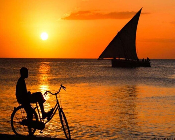 A silhouette of a person on a bicycle against a sunset with a sailboat in the water. 5
