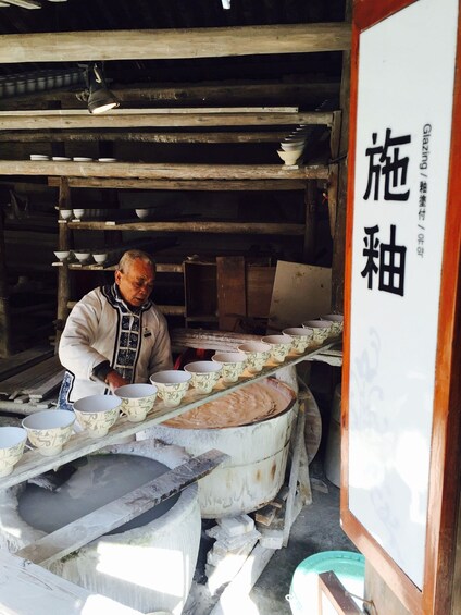 A pottery workshop with a man working on pottery wheels, shelves with bowls, and a sign with Chinese characters. 4