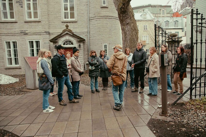 A group of people standing on a paved walkway in front of a historic building with a stone facade and a large tree. 3