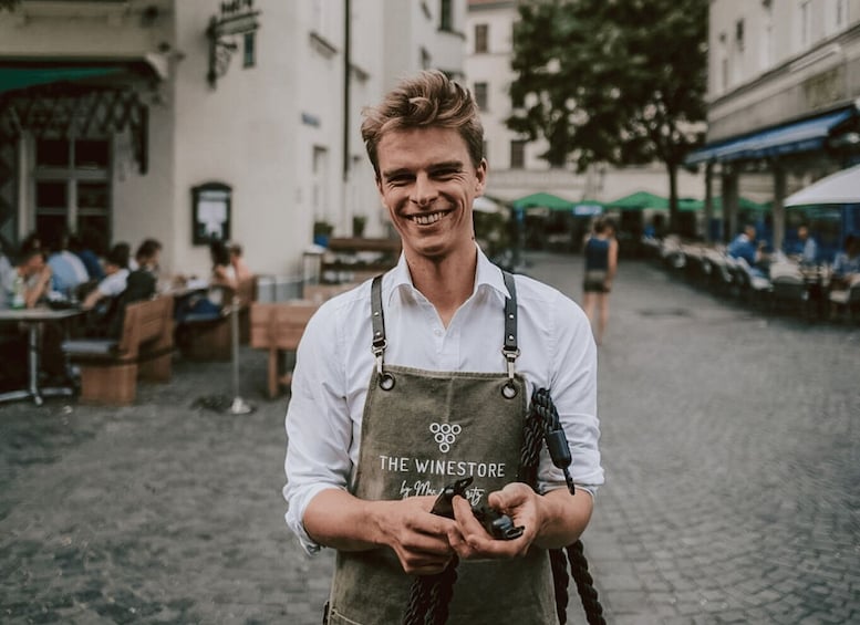 A person in a white shirt and apron with 'THE WINESTORE' text, holding a camera on a cobblestone street with outdoor seating and buildings in the background. 2
