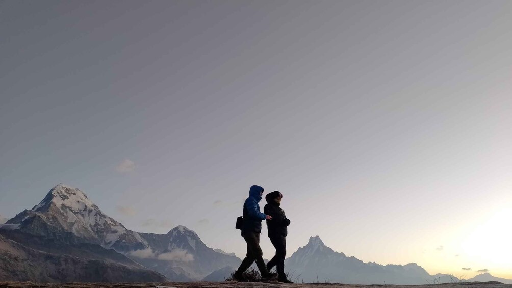 Two people walking on a mountain trail with snow-capped peaks in the background. 1